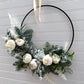 Side view of a black metal hoop wreath hanging from an ivory ribbon; decorated with lambs ear, contrasting green foliage, pieces of white fern, and small ivory peony flowers