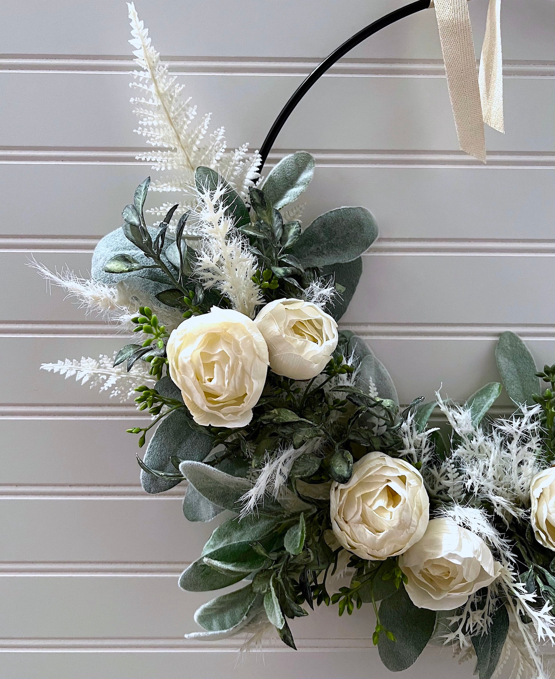 Magnified  view of a black metal hoop wreath hanging from an ivory ribbon; decorated with lambs ear, contrasting green foliage, pieces of white fern, and small ivory peony flowers